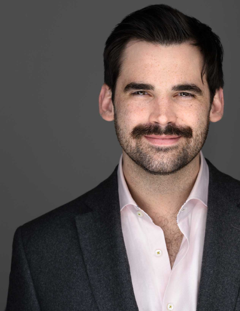 Photo of Kyle. Hes standing in front of a solid gray background. Hes wearing a white collared shirt and black suit jacket. He has short black hair and a black mustache. he is smiling