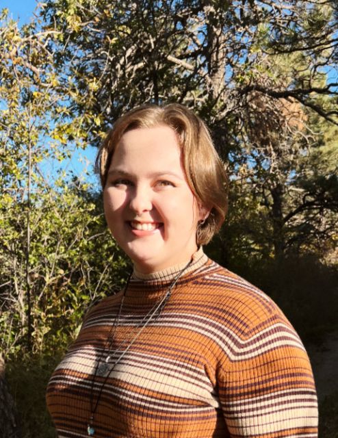 Shannon standing and smiling. She is wearing an orange and brown turtle neck top. Her hair is sandy and is cropped short. Behind her are trees