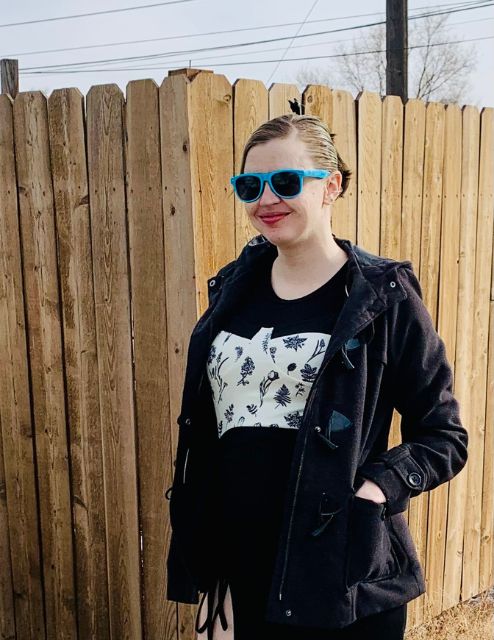 Helen is standing smiling at the camera, wearing a black outfit and blue sunglasses. She is standing outside in front of a fence with her hands in her pockets. 