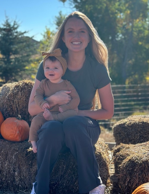 Heidi and her baby girl are in a pumpkin patch, both smiling at the camera. 
