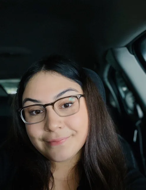 Photo of Abigail Sisneros. Dark straight hair and wearing black glasses. She is smiling